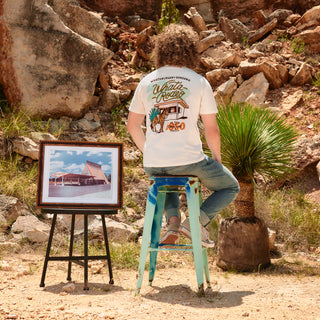 Person sitting on a stool in a desert setting with a framed picture and 'Whata Rodeo' t-shirt.
