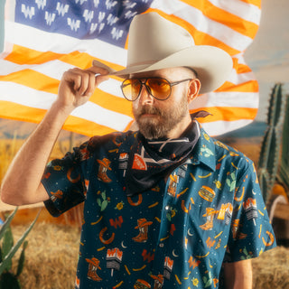 A rugged-looking man tips his cowboy hat while standing in front of an orange and white flag with Whataburger logos. He wears the Navy Collage Snap Shirt, featuring a playful Western motif, and accessorizes with amber aviator sunglasses and a navy bandana with Whataburger branding. The rustic desert setting complements the outfit’s bold Texas-inspired design.