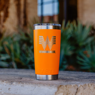 An orange 20oz Whataburger Yeti Rambler® tumbler with a stainless steel base, featuring the iconic Whataburger 'W' logo and text engraved on the surface, placed on a stone surface with a blurred natural background.