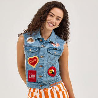 The female model is facing the camera, wearing the Wrangler® x Whataburger® Ladies Cut-Off Vest over a simple top. She poses with her hand in the pocket of her flared orange-and-white striped jeans, smiling. The vest is decorated with multiple patches inspired by Whataburger branding and Wrangler's western heritage.