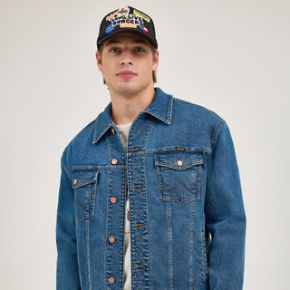 A male model wearing the classic blue denim jacket with copper buttons, contrast stitching, and chest pockets. He pairs it with a white t-shirt and a black cap featuring embroidered patches.