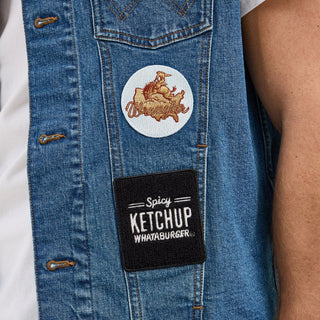 Close-up of patches on the Wrangler® x Whataburger® Men's Cut-Off Vest – A detailed view of the Wrangler® x Whataburger® Men's Cut-Off Vest, featuring a Wrangler® Texas-shaped embroidered patch and a Whataburger® Spicy Ketchup patch on the lower side.