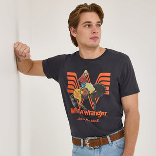 Front View – A male model wearing the Wrangler® x Whataburger® Long Live Burgers T-Shirt in faded black, featuring a bold orange and white Western-themed graphic of a cowboy riding a bucking bronco in front of the Whataburger "W" logo. He is styled with a brown leather belt and light wash jeans.