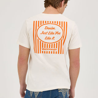 A male model wears the Wrangler® x Whataburger® Just Like You Like It T-Shirt in off-white. The t-shirt features a small "WHATAWrangler" logo in orange on the left chest. He pairs it with blue denim jeans and classic sneakers, standing in a relaxed pose.