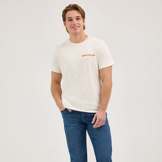 A male model wears the Wrangler® x Whataburger® Just Like You Like It T-Shirt in off-white. The t-shirt features a small "WHATAWrangler" logo in orange on the left chest. He pairs it with blue denim jeans and classic sneakers, standing in a relaxed pose.