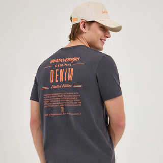 The model is turned away from the camera, showcasing the back of the black Wrangler® x Whataburger® Ingredient T-Shirt. The back design features bold orange typography reading "WHAT A Wrangler ORIGINAL DENIM Limited Edition," followed by a list of "ingredients" describing the authenticity of Wrangler’s denim and cowboy culture elements. The model wears a beige Whataburger-themed mesh trucker hat.