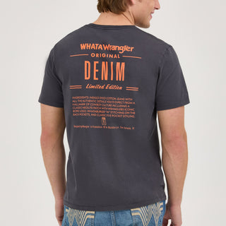 The model is turned away from the camera, showcasing the back of the black Wrangler® x Whataburger® Ingredient T-Shirt. The back design features bold orange typography reading "WHAT A Wrangler ORIGINAL DENIM Limited Edition," followed by a list of "ingredients" describing the authenticity of Wrangler’s denim and cowboy culture elements. The model wears a beige Whataburger-themed mesh trucker hat.