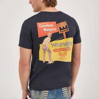 A model wearing the Wrangler® x Whataburger® Cowboy Burgers Black T-Shirt, standing at an angle to showcase the back design. The back graphic features vintage-inspired Wrangler and Whataburger signage, including a cowboy, "Cowboy Burgers" text, and the Flying W logo.