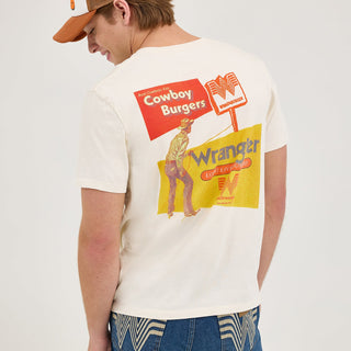 A male model wearing the Wrangler® x Whataburger® Cowboy Burgers White T-Shirt, paired with classic blue jeans and brown boots. The front of the shirt features a small Wrangler® and Whataburger® co-branded logo with "Long Live Burgers" in orange text.