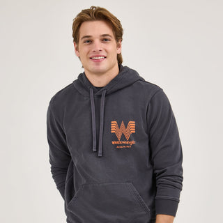 A male model wearing the Wrangler® x Whataburger® Faded Black Hoodie, smiling while standing with his hands in his hoodie pocket. The orange Flying W logo and "WHATAWrangler" branding are visible on the chest.