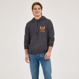 A full-body shot of a male model wearing the Wrangler® x Whataburger® Faded Black Hoodie, paired with blue jeans and cowboy boots. The hoodie has a relaxed fit, with the Flying W logo in orange on the chest.