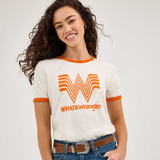 A woman with curly hair models the Wrangler® x Whataburger® Women's Ringer Tee, a cream-colored shirt with orange trim on the collar and sleeves. The front of the shirt features the Whataburger "W" logo in orange with "WHATAWrangler" written below it. She pairs it with blue denim jeans and a brown belt.