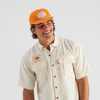 Man wearing a beige shirt and orange cap with branding on a light gray background