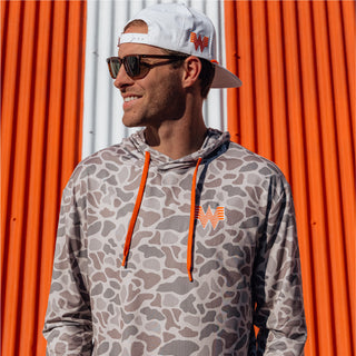 Man wearing a camo Whataburger hoodie and white Whataburger cap, smiling while standing in front of striped background.