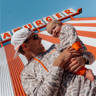 Man holding a smiling baby, both in matching gray camo Whataburger outfits, with Whataburger signage in the background.