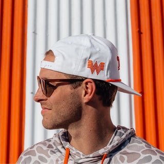 Man in a camo hoodie and white Whataburger cap worn backwards, standing against a striped background.