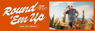 Western-themed web banner promoting new spring styles. The bold, cursive text 'Round 'Em Up' appears on an orange background, accompanied by 'New Spring Styles Are Here' in a smaller oval label. Below, a call-to-action reads 'Shop New Arrivals' with an arrow pointing toward a photograph of a bearded man in a cowboy hat, aviator sunglasses, and a red bandana. He wears the Navy Collage Snap Shirt and Swim Shorts, standing in a desert scene with cacti, hay bales, and a wooden fence.