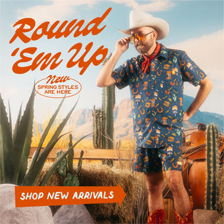 Square web banner with a Western theme promoting new spring styles. The top-left corner features the stylized text 'Round 'Em Up' in orange script, with 'New Spring Styles Are Here' in an oval beside it. Below, an orange arrow-shaped button reads 'Shop New Arrivals.' The right side displays a bearded man dressed in the Navy Collage Snap Shirt and Swim Shorts, tipping his cowboy hat while standing in a desert-inspired setting with hay bales, cacti, and a wooden fence.