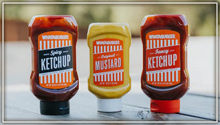 View Grocery Collection - Spicy Ketchup, Original Mustard, and Fancy Ketchup lined up in a row on a picnic table. Reads from our kitchen to yours.