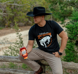 view cowboy sitting on fence in cowboy hat holding a bottle of spicy ketchup with cholula®, wearing ketchup with a kick tee from whataburger
