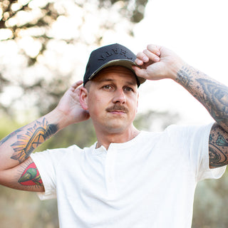 Man in a white shirt adjusts his black Whataburger x True Brvnd hat with a camo-patterned brim, standing outdoors.