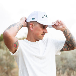 Man in a white shirt adjusts his white Whataburger x True Brvnd hat with a black embroidered "WHATA" on the front, standing outdoors.