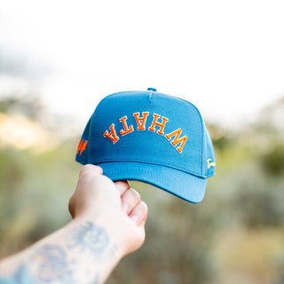 Hand holds up a blue Whataburger x True Brvnd cap against a natural outdoor background. The cap features bold orange and white embroidered lettering spelling an upside down, curved 'WHATA' across the front, with subtle branding details on the sides.