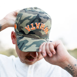 Close-up of a man adjusting camo Whataburger x True Brvnd cap. The brim partially shields his face, highlighting the orange and white embroidered 'WHATA' embroidery on the front.