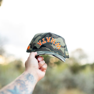 Hand holds up a camo-print Whataburger x True Brvnd cap against a natural outdoor background. The cap features orange and white embroidered lettering spelling an upside down, curved 'WHATA' across the front, with subtle branding details on the sides.