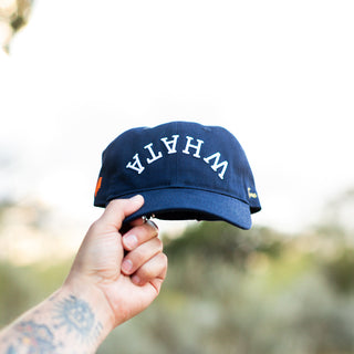 Hand holds up a navy blue Whataburger x True Brvnd dad hat against a natural outdoor background. The cap features white embroidered lettering spelling an upside down, curved 'WHATA' across the front, with subtle branding details on the sides.