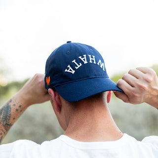 Close-up of a man adjusting navy blue Whataburger x True Brvnd dad hat sitting backwards on his head, highlighting white embroidered 'WHATA' embroidery on the front.