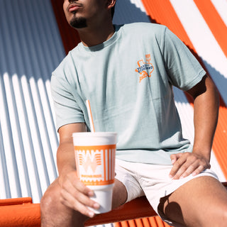 Person wearing a light blue t-shirt with an orange logo, holding a cup with an orange straw, sitting on an orange chair against a striped wall.