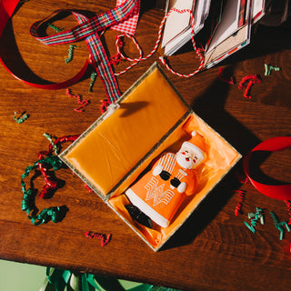 Small Santa Claus ornament in an open box on a wooden surface with ribbons and decorations.