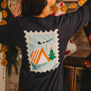 Person wearing a navy blue long-sleeve shirt with a festive design of Santa Claus, a campfire, and a Christmas tree on the back.