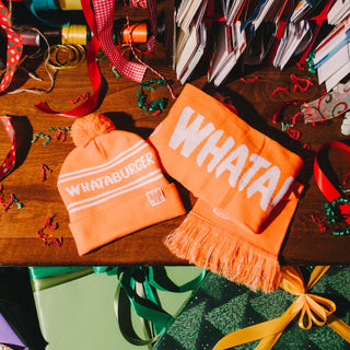 Orange beanie and scarf with 'WHATA!' text on a wooden table with festive decorations.