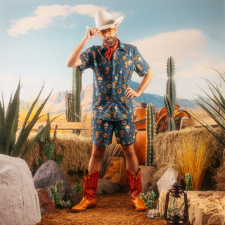 A bearded man dressed in a Western-inspired outfit stands in a desert-themed set. He wears the Navy Collage Snap Shirt and matching Navy Collage Swim Shorts, adorned with icons like cowboy boots, cacti, Whataburger signage, and horseshoes. He pairs the look with a white cowboy hat, red bandana, orange-tinted aviator glasses, and red cowboy boots. The backdrop features hay bales, cacti, and a wooden fence, evoking a Southwestern atmosphere.