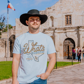 A man in a black cowboy hat stands in front of the Alamo, wearing a light blue Whataburger-themed t-shirt featuring a rope-style "Whataburger" text inside the shape of Texas. The shirt also includes the phrase "Just how Y'ALL like it" and a "75 years" badge.