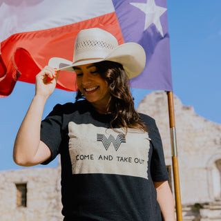 A smiling woman in a white cowboy hat poses in front of the Texas flag and the Alamo, wearing a black Whataburger t-shirt with a distressed design. The shirt features the Whataburger "W" logo and the phrase "COME AND TAKE OUT" in bold text.