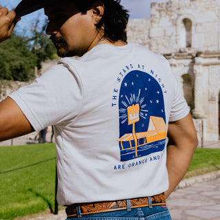 A man wearing a white t-shirt with a colorful back design stands in front of the Alamo, tipping his black cowboy hat. The shirt showcases an illustration of a Whataburger restaurant with a neon-style glow and the phrase "THE STARS AT NIGHT ARE ORANGE AND WHITE."