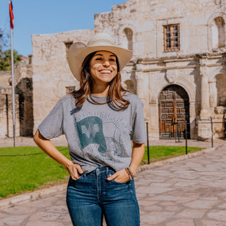 A cheerful woman in a white cowboy hat stands in front of the Alamo, wearing a gray Whataburger t-shirt. The shirt design features an illustrated Texas flag waving behind the Whataburger "W" logo, with the phrase "The Original"