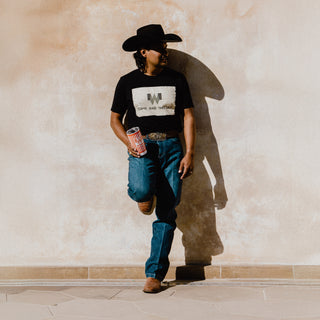 A man leans against a beige stucco wall, casting a strong shadow in the sunlight. He wears a black "COME AND TAKE OUT" Whataburger t-shirt, blue jeans, cowboy boots, and a black cowboy hat. He holds a Whataburger-striped tumbler in his hand.