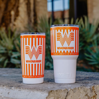 Orange and white striped 20oz and 30 oz Whataburger Yeti Rambler® tumblers with a stainless steel base, featuring the iconic Whataburger 'W' logo and text engraved on the surface, placed on a stone surface with a blurred natural background.