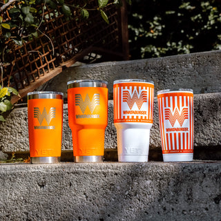 Four Whataburger-themed Yeti tumblers in orange and white designs are displayed on concrete steps, with greenery in the background.