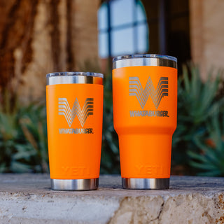 Orange 30oz and 20oz Whataburger Yeti Rambler® tumblers with a stainless steel base, featuring the iconic Whataburger 'W' logo and text engraved on the surface, placed on a stone surface with a blurred natural background.