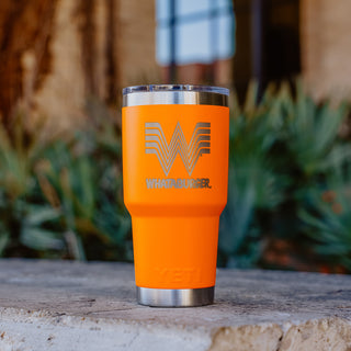 An orange 30oz Whataburger Yeti Rambler® tumbler with a stainless steel base, featuring the iconic Whataburger 'W' logo and text engraved on the surface, placed on a stone surface with a blurred natural background.