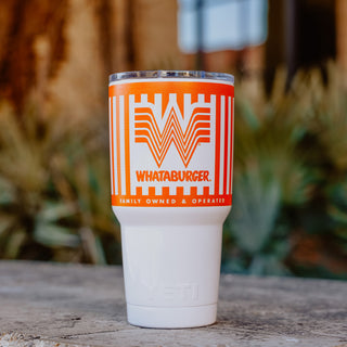 An orange and white striped 30oz Whataburger Yeti Rambler® tumbler with a stainless steel base, featuring the iconic Whataburger 'W' logo and text engraved on the surface, placed on a stone surface with a blurred natural background.