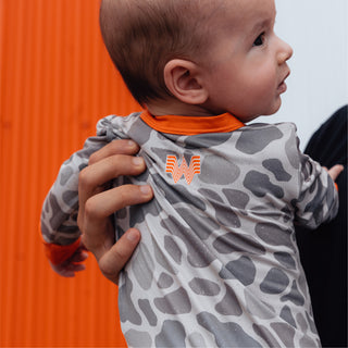 Baby held up in front of orange and white striped building wearing WHATA Classic Deer Camo Baby Zip Up.