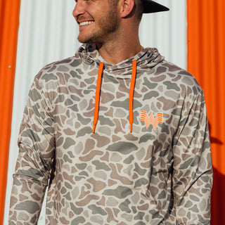 A man wearing a Classic Deer Camo  performance hoodie with orange drawstrings and a matching Whataburger chest logo, photographed against a white and orange corrugated wall.