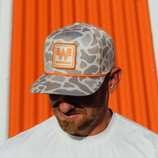 A man wearing a Classic Deer Camo cap with bright orange accents, set against a vibrant orange background.