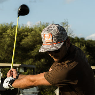 A man wearing a brown polo shirt and a Classic Deer Camo cap with an orange Whataburger logo patch is captured mid-swing on a golf course.